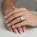 An engagement ring featuring a rectangular radiant-cut diamond center stone in a four-prong setting, with a pavé band set with small round diamonds, shown on a reflective light gray surface.